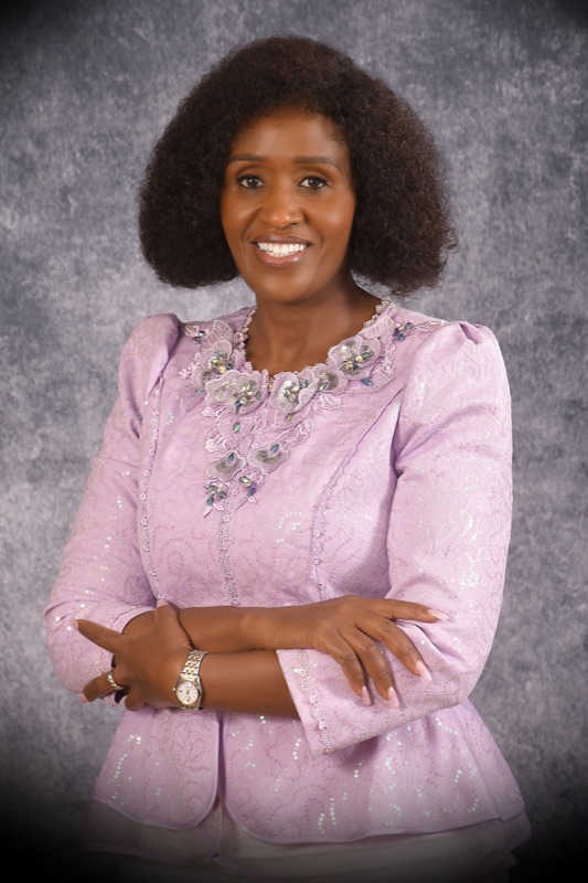 Anne Musau smiling warmly, pink blouse against a grey studio backdrop