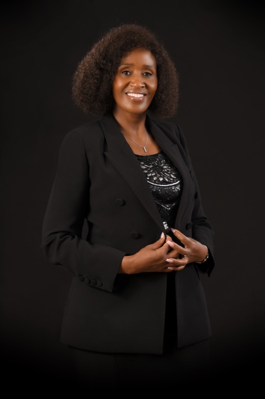 Anne Musau in a black suit, hands clasped, charcoal backdrop